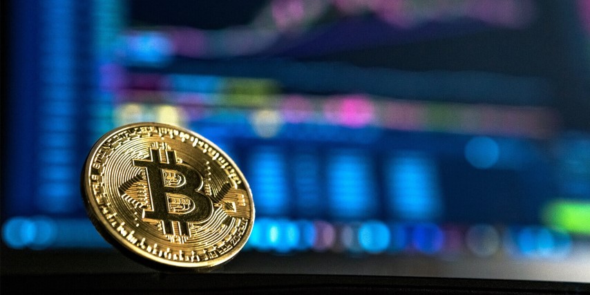A bitcoin in front of a laptop display of a cryptocurrency trading portal.